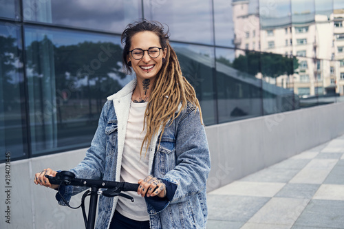 Valokuva Happy smiling woman with tattooes and dreadlocks is driving electro scooter on the street