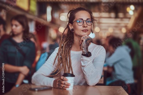 Beautiful cheerful girl with tattooes and dreadlocks is sitting at food court while drinking coffee. © Fxquadro