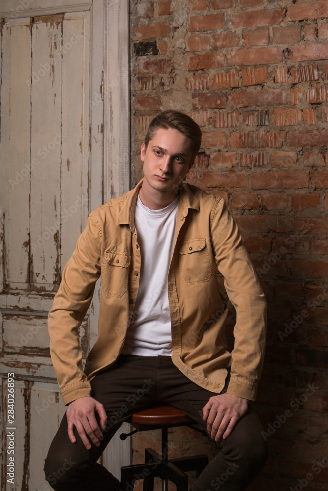 Sad and thoughtful young guy sitting on a chair. In the background is an old brick wall. Beautiful, expressive portrait made in the Studio in the loft design. Sad and thoughtful young guy sitting on a chair. In the background is an old brick wall. Beautiful, expressive portrait made in the Studio in the loft design.