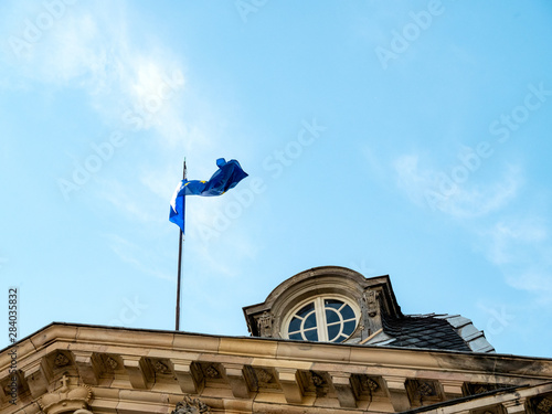 European Union blue flag waving calmly on the rooftop of Palais Rohan in Strasbourg with beautiful ornate window and clear blue sky in background