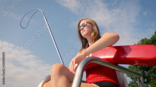 Low angle of a female lifeguard on a stand watching over swimmers at a public pool. The teen blows her whistle while working a summer job lifeguarding. Action shot represents employment and safety. photo