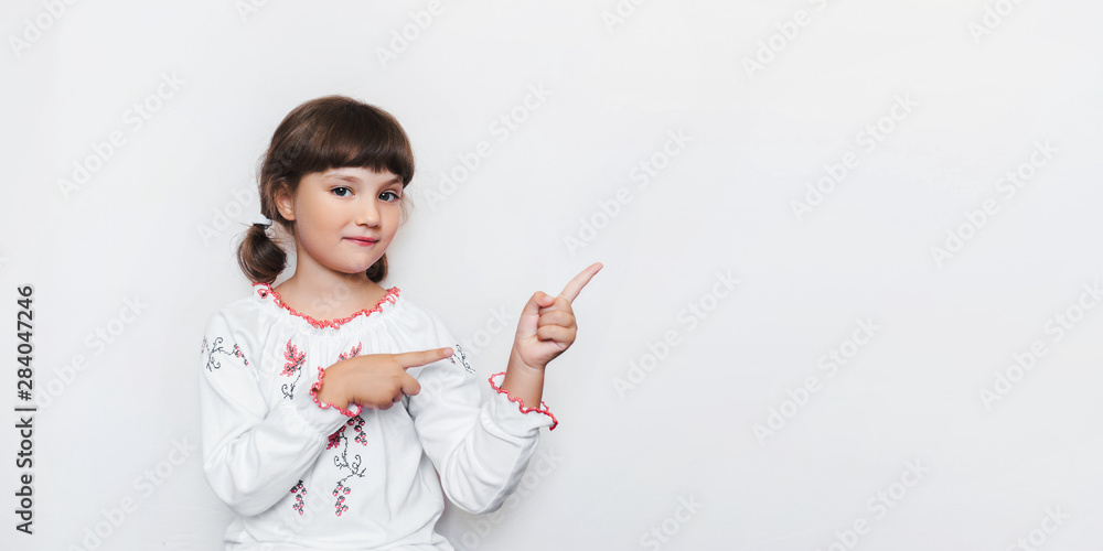 A little Ukrainian girl in a white patterned embroidery on the background of the wall shows with index fingers towards empty space. An emotional little and patriotic child. Creative photography. Copy A little Ukrainian girl in a white patterned embroidery on the background of the wall shows with index fingers towards empty space. An emotional little and patriotic child. Creative photography. Copy