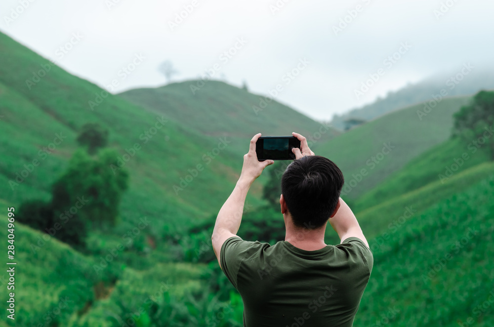 A male person using mobile phone taking a photo of greenery mountain with fog in the morning.