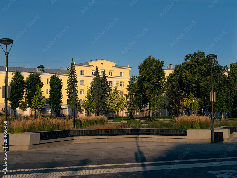 The bright dawn sun illuminates the trees and buildings in the square in the city.