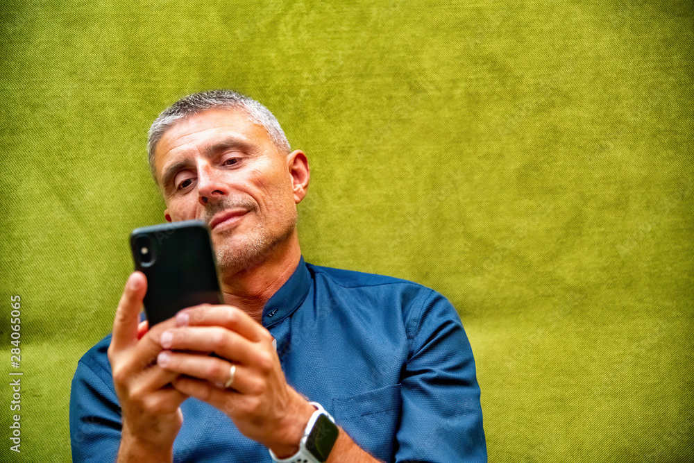 Smiling man texting on smartphone while seated on a green sofa Smiling man texting on smartphone while seated on a green sofa