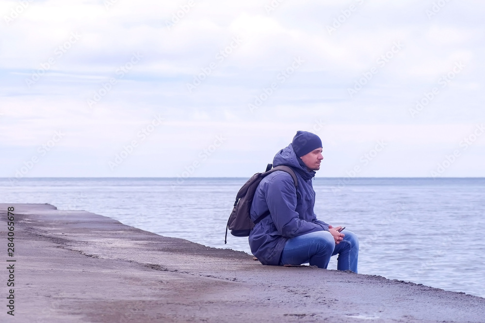 Young man traveller with backpack and smartphone in hands wears in jacket and hat at sea. Man tourist comes to old sea waterfront sits and looks at sea in winter. Overcast day at se. Young man traveller with backpack and smartphone in hands wears in jacket and hat at sea. Man tourist comes to old sea waterfront sits and looks at sea in winter. Overcast day at se.