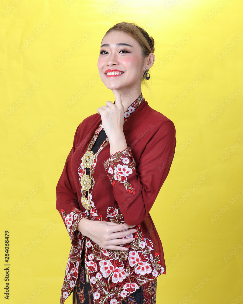 Asian woman traditional red maroon kebaya sharong on yellow paper background posing stand happy finger on chin Asian woman traditional red maroon kebaya sharong on yellow paper background posing stand happy finger on chin