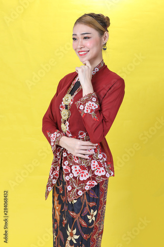 Asian woman traditional red maroon kebaya sharong on yellow paper background posing stand happy finger on chin