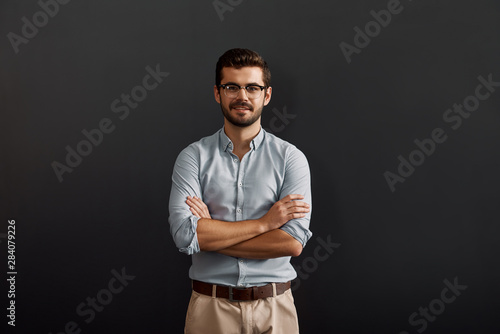 Project manager. Confident and young bearded man looking at camera with smile and keeping arms crossed while standing against dark background