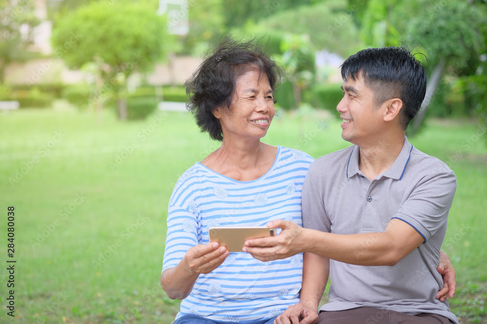 Middle-aged Asian mother and son looking at each other and looking at a smartphone with a smile and being happy at the park Is an impressive warmth Middle-aged Asian mother and son looking at each other and looking at a smartphone with a smile and being happy at the park Is an impressive warmth