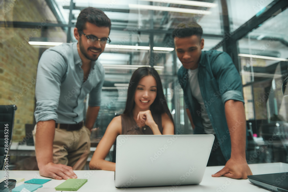 Using modern technologies. Group of three young and cheerful employees looking at screen of laptop and smiling while working in modern office