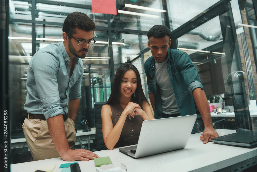Project team. Group of three young employees looking at screen of laptop and discussing new project while working in modern office Project team. Group of three young employees looking at screen of laptop and discussing new project while working in modern office