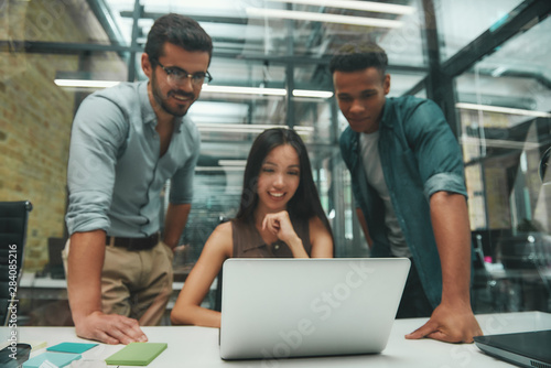 Using modern technologies. Group of three young and cheerful employees looking at screen of laptop and smiling while working in modern office