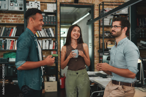 Fresh office news. Young smiling colleagues in casual wear holding cups and talking about something while standing in modern office © Friends Stock