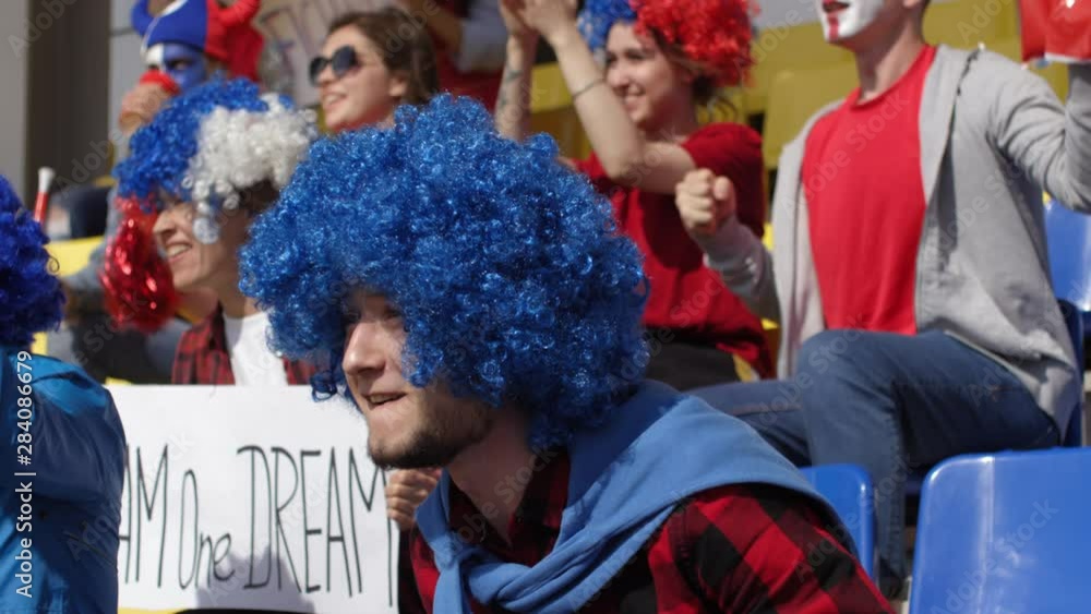 Tilting shot of excited young Caucasian football fan in bright blue wig anxiously watching match, shouting, cheering, jumping up from seat and blowing whistle to celebrate goal with other supporters Tilting shot of excited young Caucasian football fan in bright blue wig anxiously watching match, shouting, cheering, jumping up from seat and blowing whistle to celebrate goal with other supporters