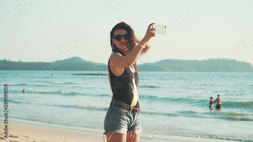 Pretty young woman in sunglasses and swimsuit takes selfie on beautiful sandy beach by sea. Happy smiling girl pulls her hair, spinning and taking pictures of herself with smartphone. Slow motion.