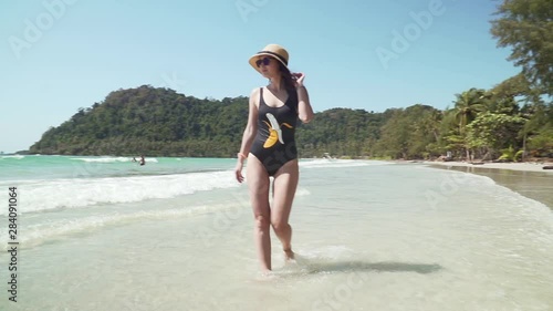 Young caucasian woman walks along sandy beach by the sea. Girl in swimsuit and straw hat enjoys vacation on paradise tropical island with sandy beach, calm turquoise sea and green trees on the shore.