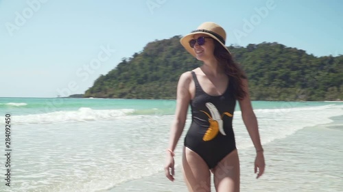 Young caucasian woman walks along sandy beach by the sea. Girl in swimsuit and straw hat enjoys vacation on paradise tropical island with sandy beach, calm turquoise sea and green trees on the shore.
