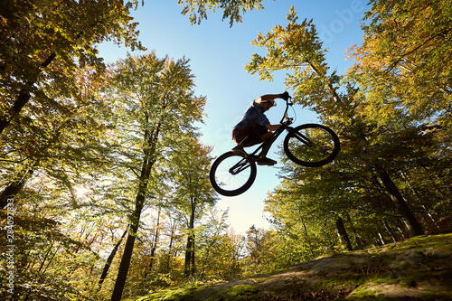 Young male bicyclist jumping on trial bicycle on big boulder, professional sportsman making acrobatic stunt in the forest on sunny day. Concept of extreme sport active lifestyle