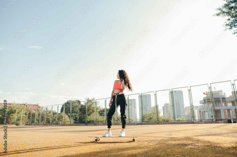 Stylish girl standing on a longboard on an old tennis court and posing at the camera, wearing stylish street clothes, looking away.Attractive hispanic girl rides longboard on street. Street style Stylish girl standing on a longboard on an old tennis court and posing at the camera, wearing stylish street clothes, looking away.Attractive hispanic girl rides longboard on street. Street style