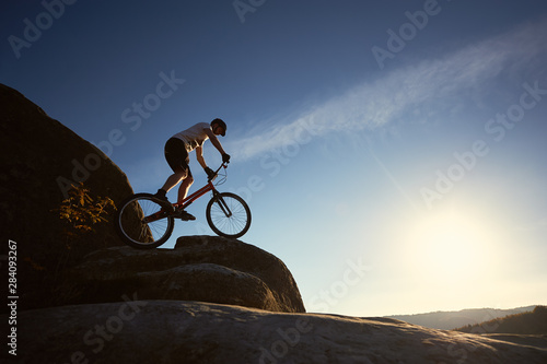 Silhouette of man cyclist balancing on trial bicycle, making acrobatic trick on top of mountain on summer sunny evening, blue sky and sunset on background. Concept of extreme sport active lifestyle