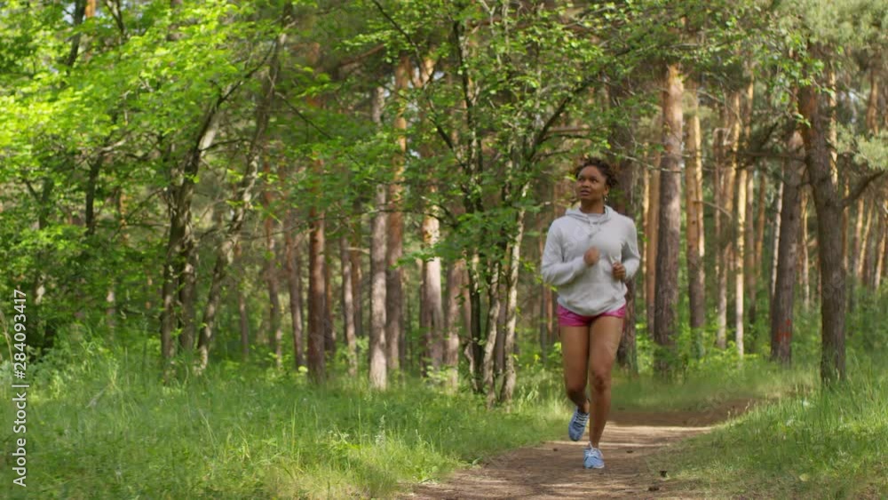 Full tracking shot of slim young Afro-American girl with braided hair, wearing white hoodie and pink shorts, running vigorously along footpath in summer forest on sunny morning