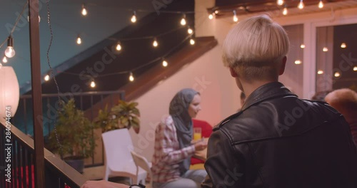 Closeup portrait of young blonde female in glasses standing happily alone and joining diverse multiracial group of friendsat party with fairy lights photo