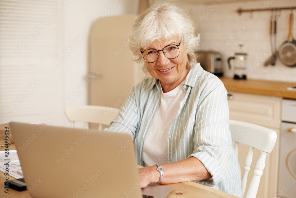 Attractive serious self employed woman pensioner working distantly from home, sitting in kitchen in front of open portable computer, wearing eyeglasses. People, age, job and occupation concept Attractive serious self employed woman pensioner working distantly from home, sitting in kitchen in front of open portable computer, wearing eyeglasses. People, age, job and occupation concept