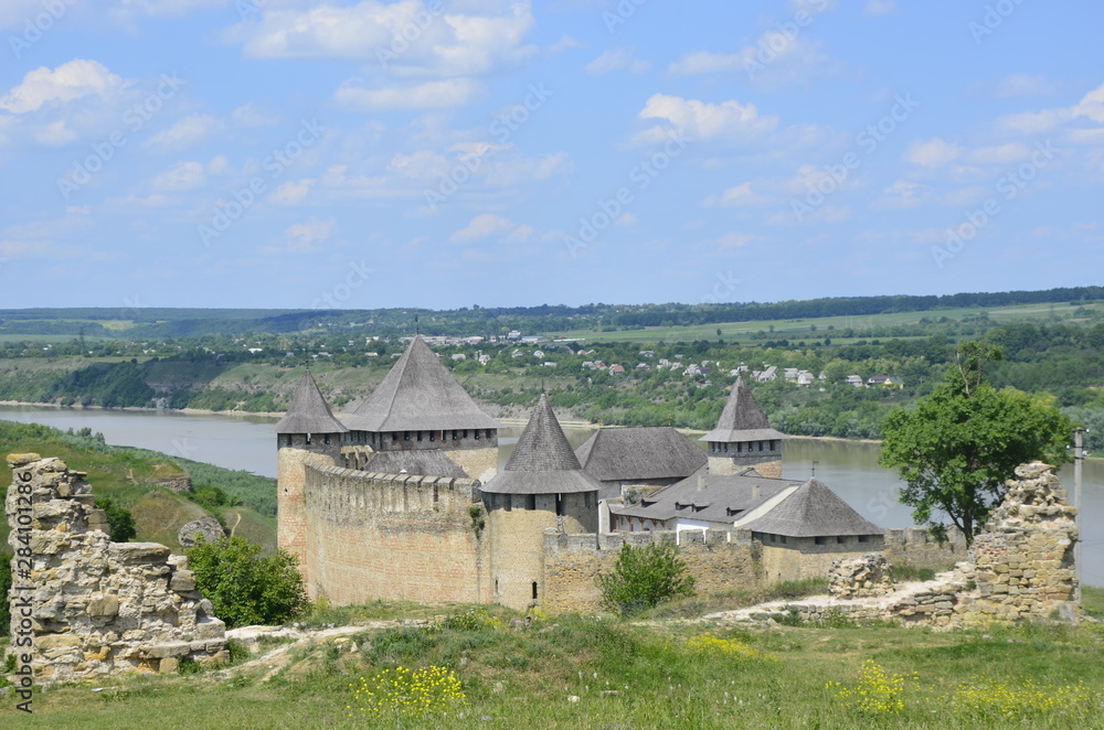 Obraz premium Magic Khotun fortress in Ukraine against the background of a blue-haired sky and white clouds, and green grass, trees and Dniester river
