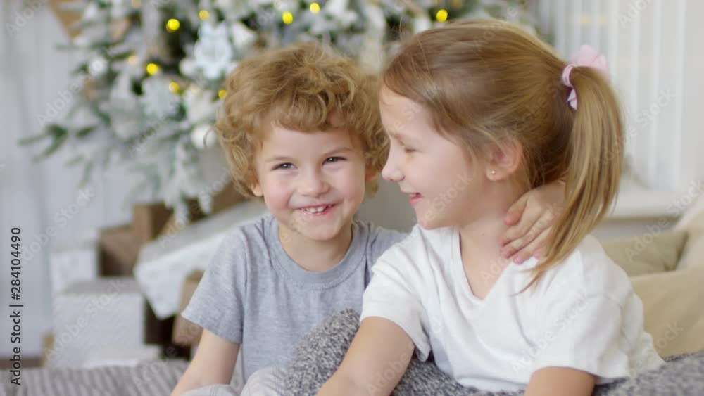 Happy little brother and sister smiling, embracing and posing for camera on bed in room decorated with Christmas tree and fairy lights Happy little brother and sister smiling, embracing and posing for camera on bed in room decorated with Christmas tree and fairy lights