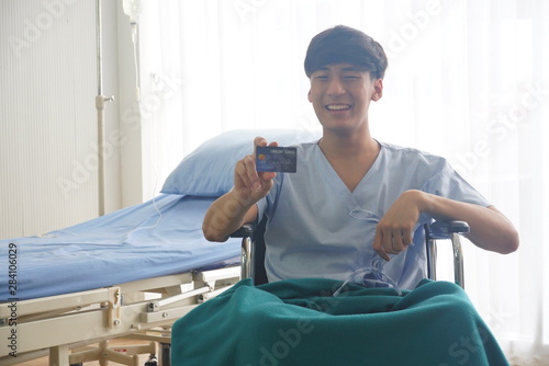 Asian patient handsome man sit on a wheelchair and shows the credit card with confidence with sunlight from the window in hospital room.
