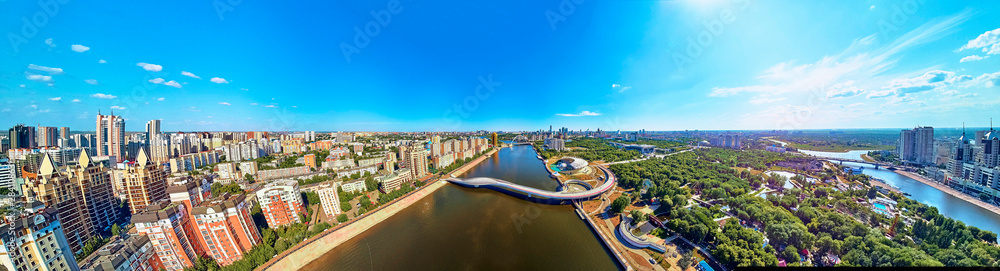 Obraz premium NUR-SULTAN, KAZAKHSTAN - July 30, 2019: Beautiful panoramic aerial drone view to Ishim River Embankment and Nursultan (Astana) city center with skyscrapers and modern pedestrian bridge