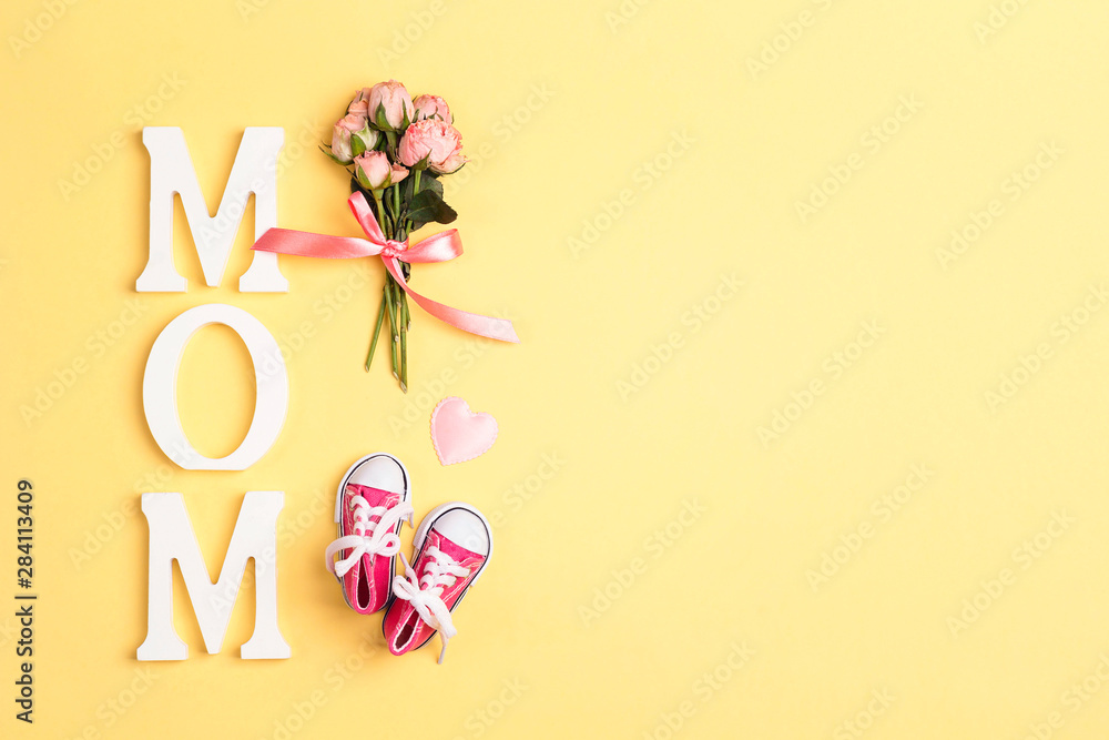 Word mom with pink baby girl sneakers and small bouquet of roses on a yellow background.