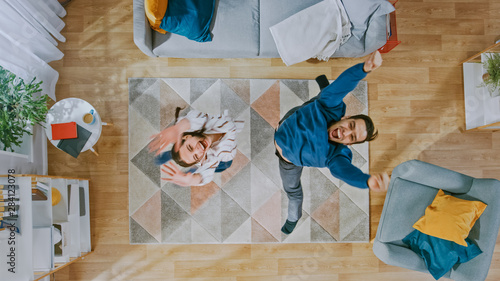 Young Happy Couple is Energetically Dancing and Jumping in an Apartment. Cozy Living Room with Modern Interior with Carpet, Sofa, Chair, Coffe Table, Book Shelf, Plant and Wooden Floor. Top Down.