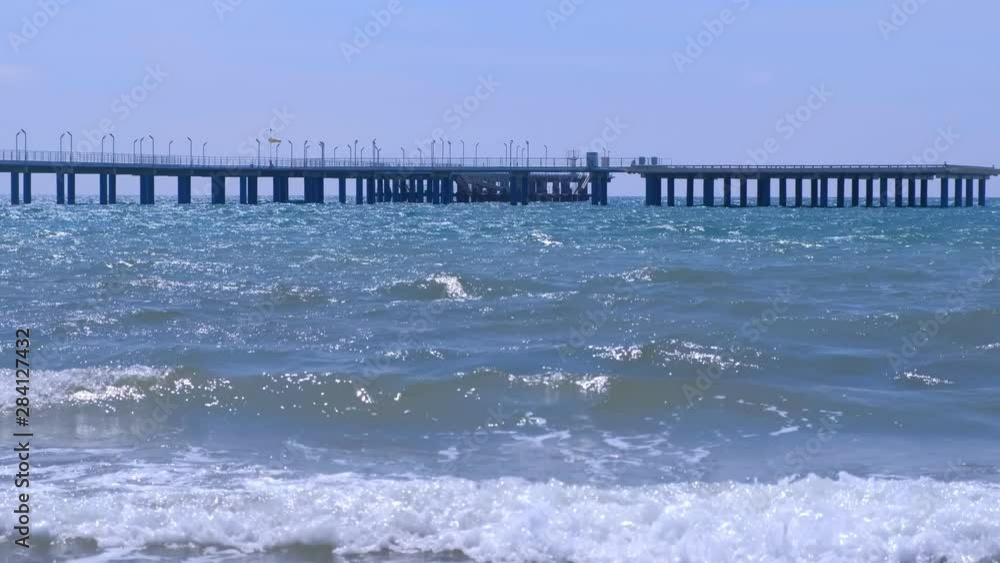 Big long pier with streetlights, fence and helicopter platform at sea. Beautiful seascape with sea waves and clear sky at sunny windy day on sand beach. Big long pier with streetlights, fence and helicopter platform at sea. Beautiful seascape with sea waves and clear sky at sunny windy day on sand beach.