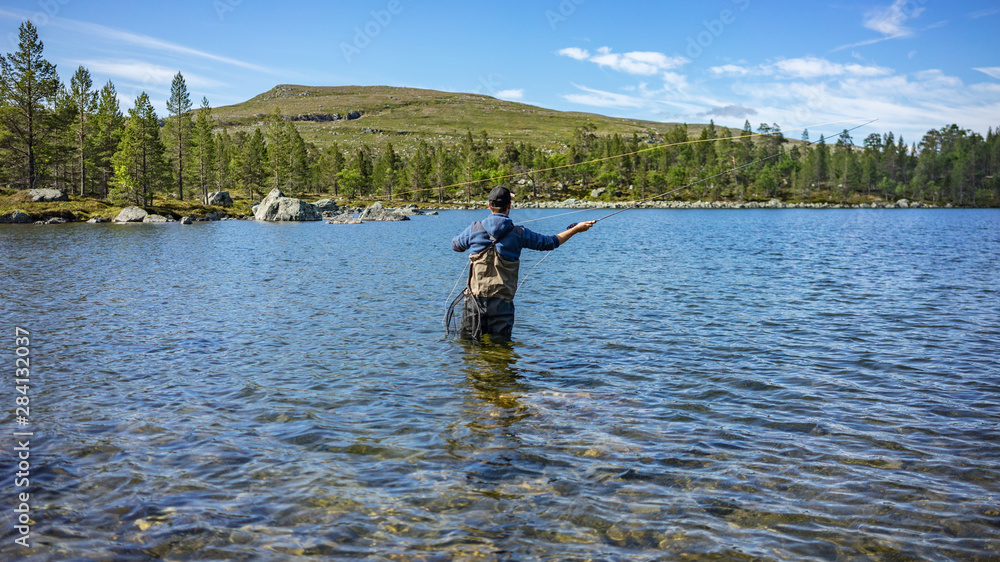 Angler mit Angel in Wathose im Wasser beim Fliegenfischen bei Sonne vor Wald und Berg bei Sonne