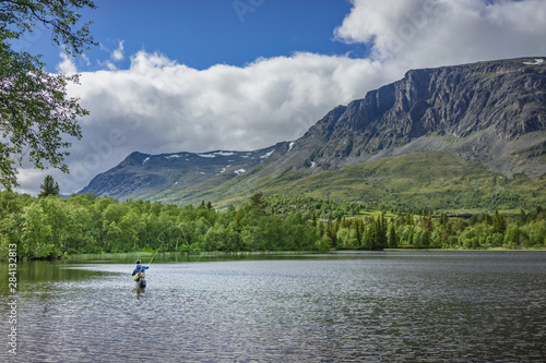 Angler beim Fliegenfischen mit Angelroute im Wasser stehend mit Wathose im See vor Bäumen und Berg © annieze