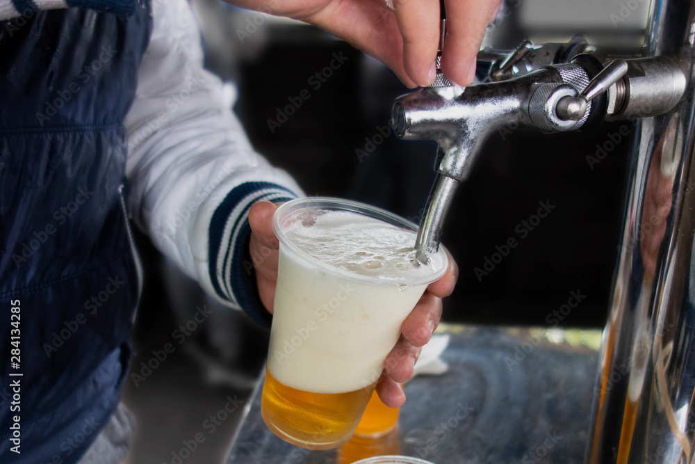 Man pours draft beer in plastic glass during picnic. Close up image with no face. Tap of movable bar counter with hands and alcohol drink