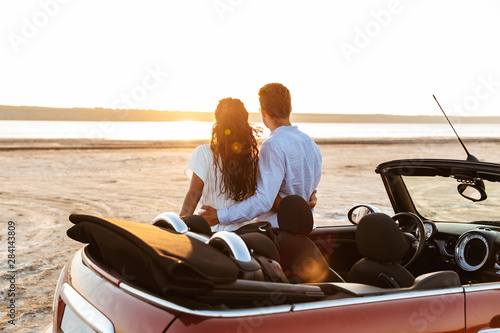 Photo from back of young multiethnic couple hugging together while standing by car on beach