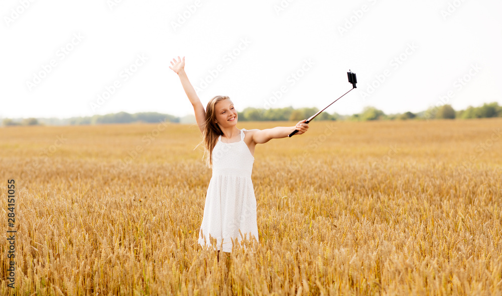 technology, summer and people concept - happy young girl in white dress taking picture by smartphone on selfie stick on cereal field technology, summer and people concept - happy young girl in white dress taking picture by smartphone on selfie stick on cereal field