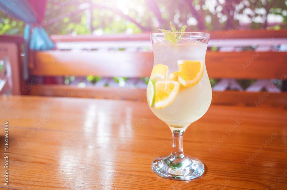 Natural citrus lemonade with slices of orange, lemon and lime in a glass on a wooden table against the background of a gazebo and bushes. Vitamin drinks concept. Bright sunshine. Natural citrus lemonade with slices of orange, lemon and lime in a glass on a wooden table against the background of a gazebo and bushes. Vitamin drinks concept. Bright sunshine.