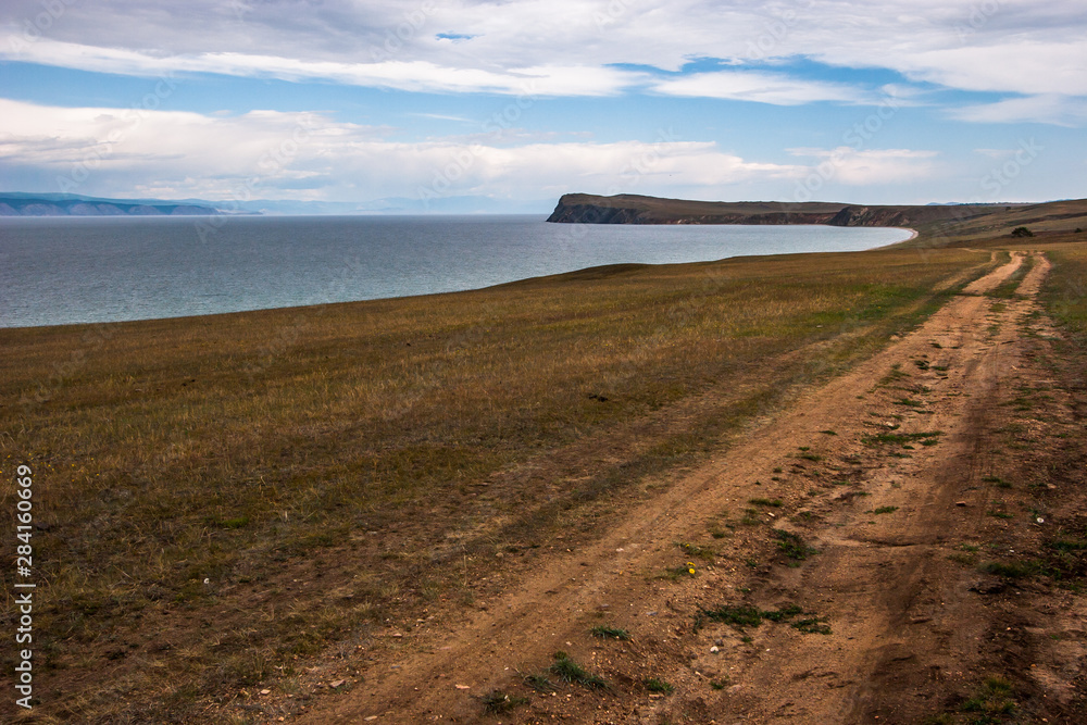 The dirt road goes along the shore of Lake Baikal to the cape. The weather is gloomy, clouds in the sky. Between the clouds blue sky. Behind the lake you can see the mountains in the haze.