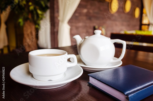 White cup of tea and teapot, notepad on a table in a cafe. The concept of tea drinking, rest, drawing up a business plan.