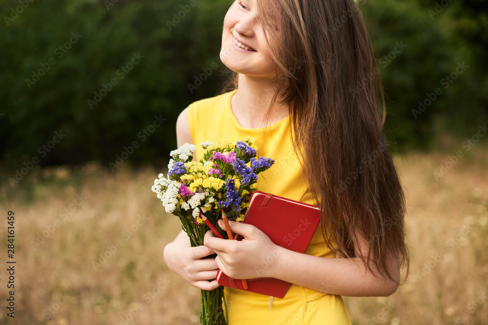 Bouquet from fields in international children's day. long-haired blond girl standing in the park with flowers, a notebook and pencils in her hands. Ready to school. Back to school Bouquet from fields in international children's day. long-haired blond girl standing in the park with flowers, a notebook and pencils in her hands. Ready to school. Back to school