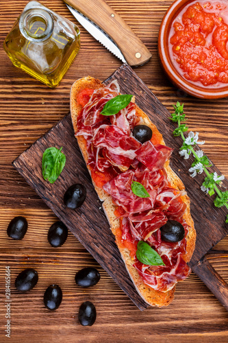 Delicious bread toast with natural tomato, extra virgin olive oil, Iberian ham, black olives and basil leaves. On wooden background.