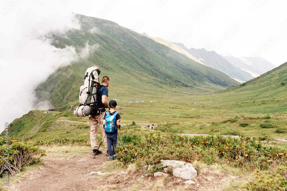 Dad and son 6-7 years old with backpacks in the mountains next to the clouds look at the campground. Parenting. Healthy athletic lifestyle.