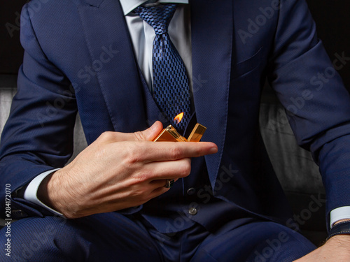 A man in a classic suit sits on a black leather sofa and shows off an expensive lighter