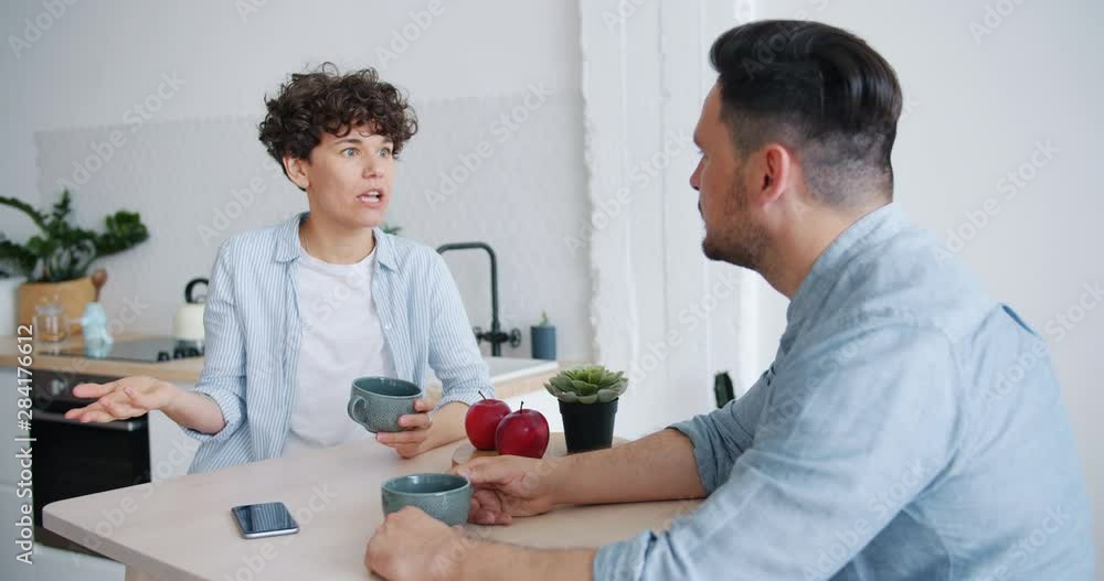 Unhappy couple man and woman are agruing sitting at table in kitchen talking with angry faces discussing marital problems. People, relationship and emotions concept. Unhappy couple man and woman are agruing sitting at table in kitchen talking with angry faces discussing marital problems. People, relationship and emotions concept.