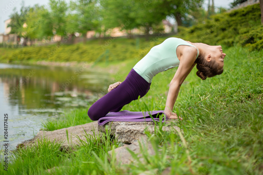 Thin brunette girl plays sports and performs beautiful and sophisticated yoga poses in a summer park. Green lush forest and the river on the background. Woman doing exercises on a yoga mat