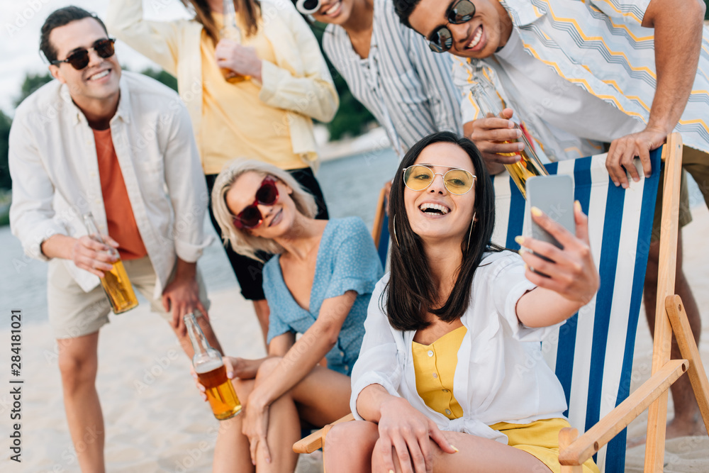 Obraz premium attractive cheerful woman taking selfie with multicultural friends having fun on beach